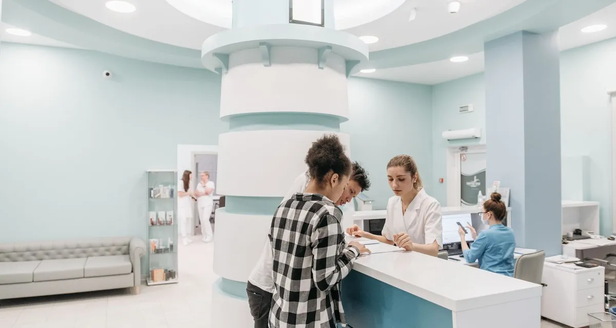 A patient completing admission paperwork at a hospital reception desk while a member of staff points to the form
