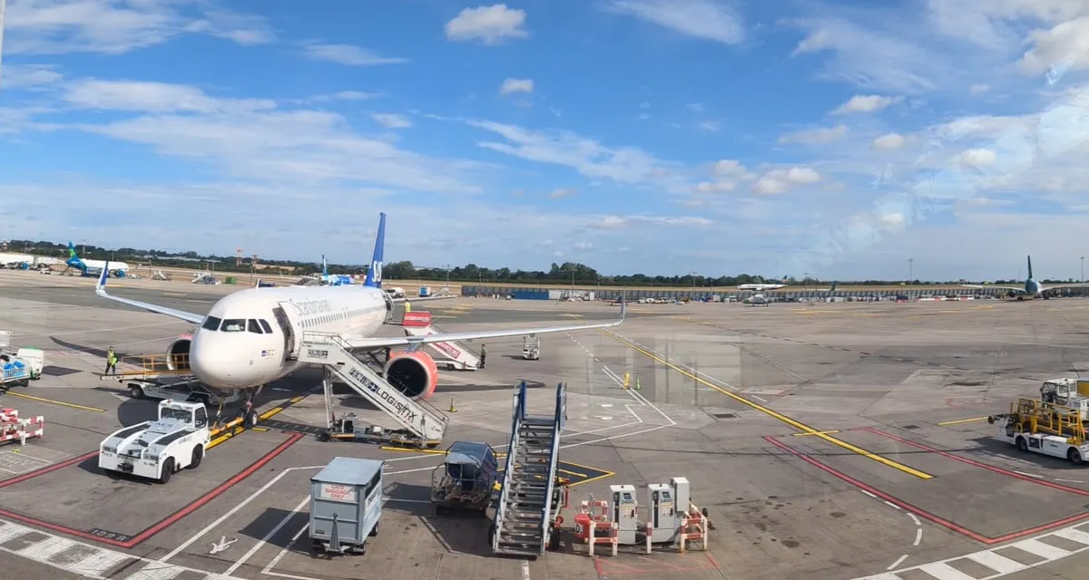 A wide view of an airport apron with a commercial airliner on stand, boarding steps in position and ground handling vehicles around it