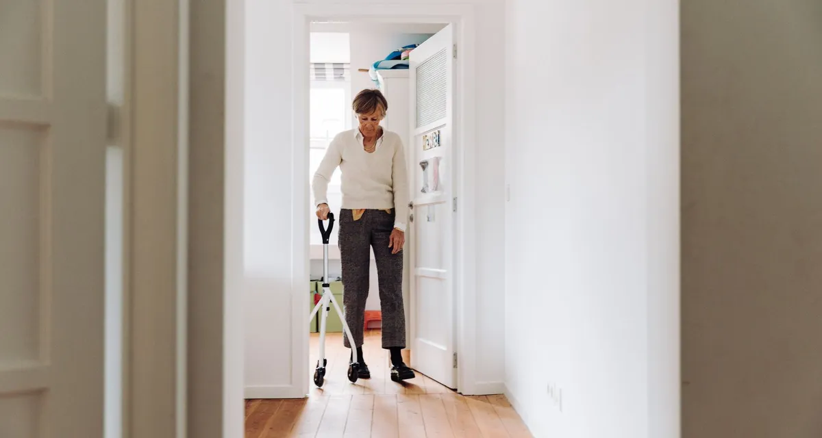 An older woman walking with a wheeled walking frame through a bright hallway at home